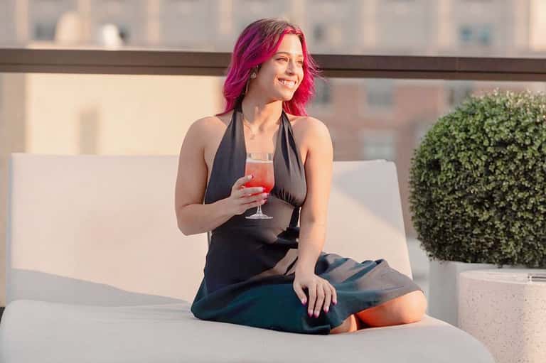 Woman with pink hair in black dress sitting on rooftop lounge with cocktail and city skyline