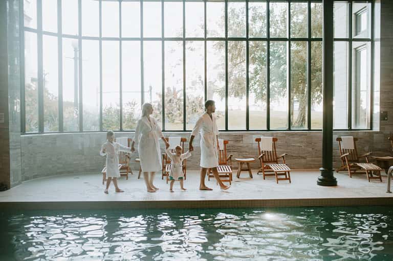 Family in white robes standing by indoor pool with large windows overlooking trees