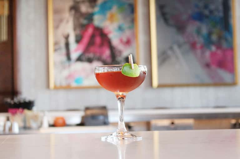 Craft cocktail with lime garnish on bar counter with blurred artwork background