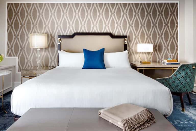 Modern hotel bedroom with neutral tones, patterned wallpaper, and blue accent pillow.