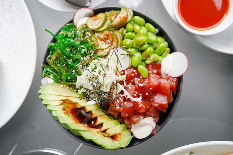 Colorful sushi bowl with seaweed, edamame, avocado, radish, salmon, and sesame seeds with red dipping sauce.