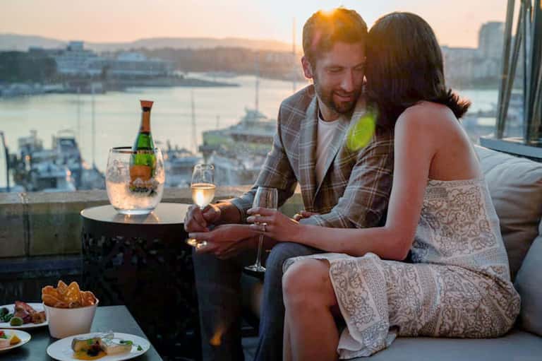 Couple enjoying champagne on high-rise balcony with city and waterfront views at sunset