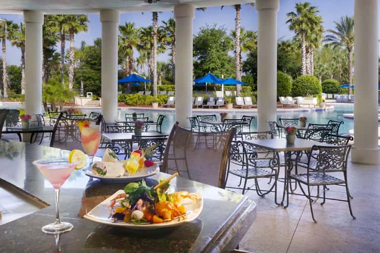 Upscale poolside dining area with palm trees, blue umbrellas, and elegant plated cuisine under a columned pavilion