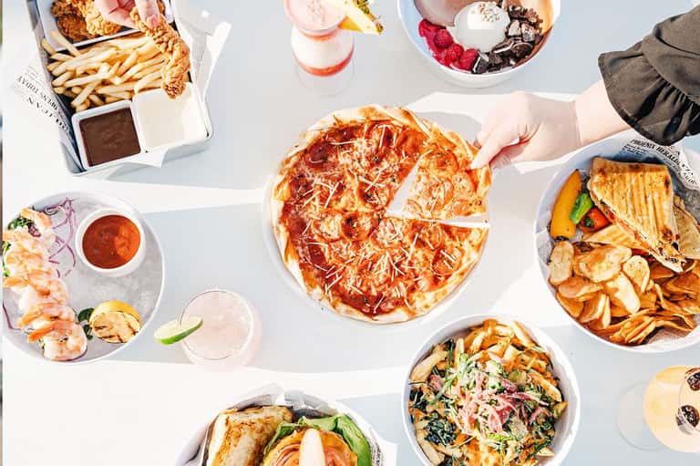 Overhead view of gourmet meal spread with pizza, seafood, drinks, and desserts