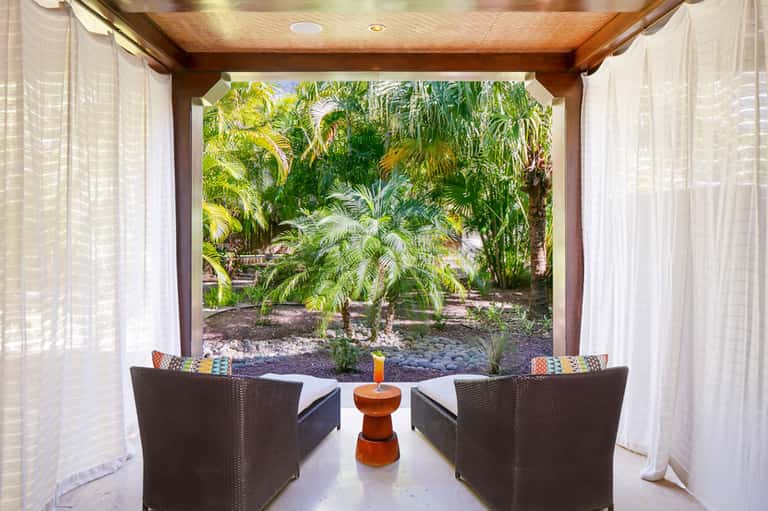 Covered outdoor seating area with wicker chairs overlooking lush tropical garden with palm trees