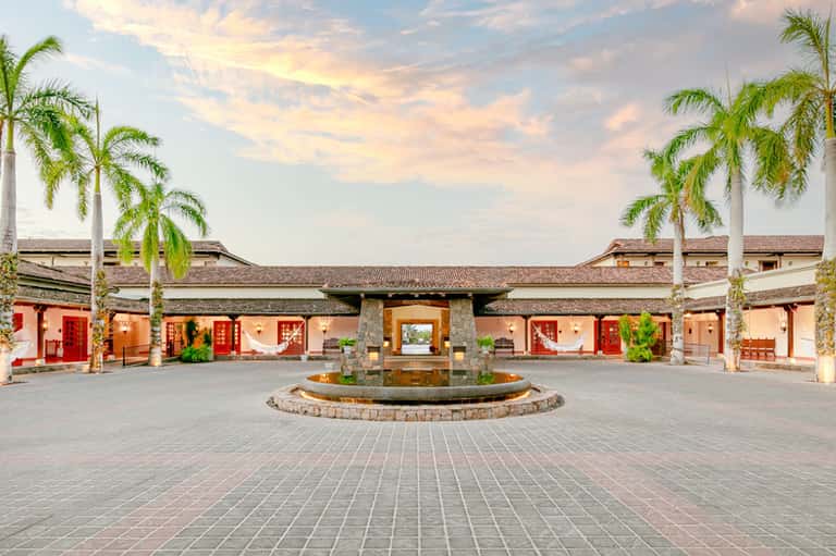 Traditional Asian-style courtyard with red doors, palm trees, and central fountain at dusk