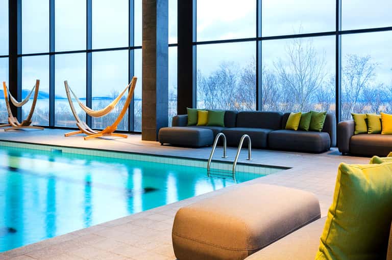Indoor pool facility with floor-to-ceiling windows overlooking winter landscape, black modern sofas with green pillows, and orange hanging chairs