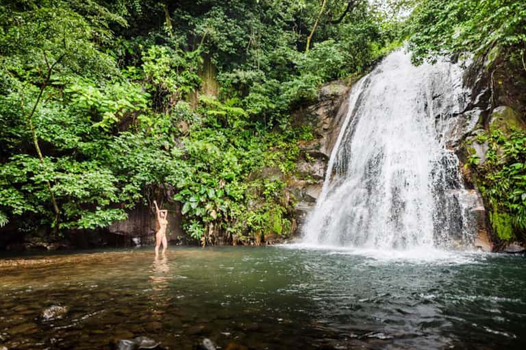 Person wading in tropical waterfall pool surrounded by lush green jungle vegetation