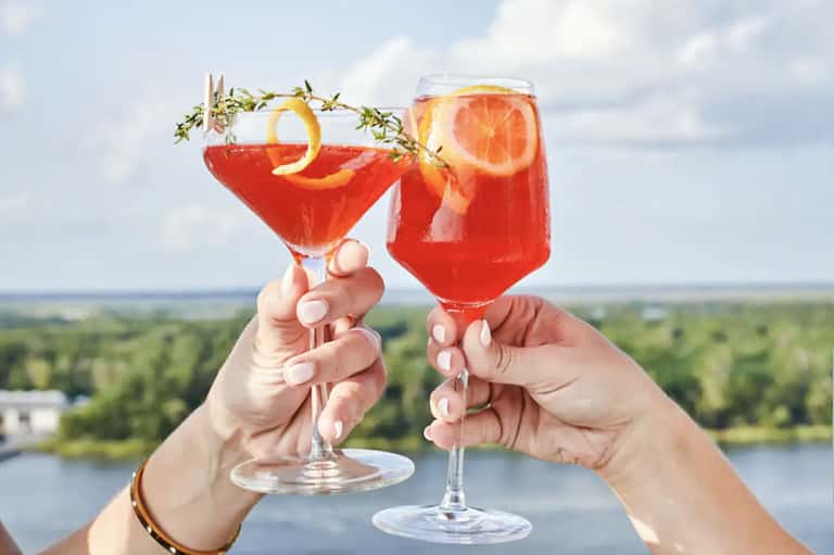 Two hands toasting with red cocktails against scenic water and forest landscape backdrop