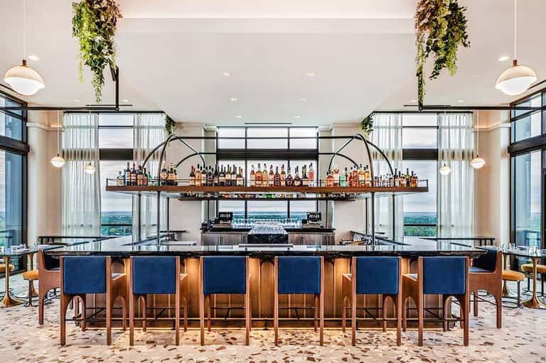 Modern beach bar with sand floor, wood bar, blue seating, hanging plants, and coastal views