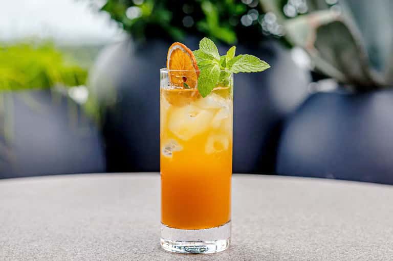 Orange cocktail with ice, mint garnish, and orange slice in tall glass by pool area