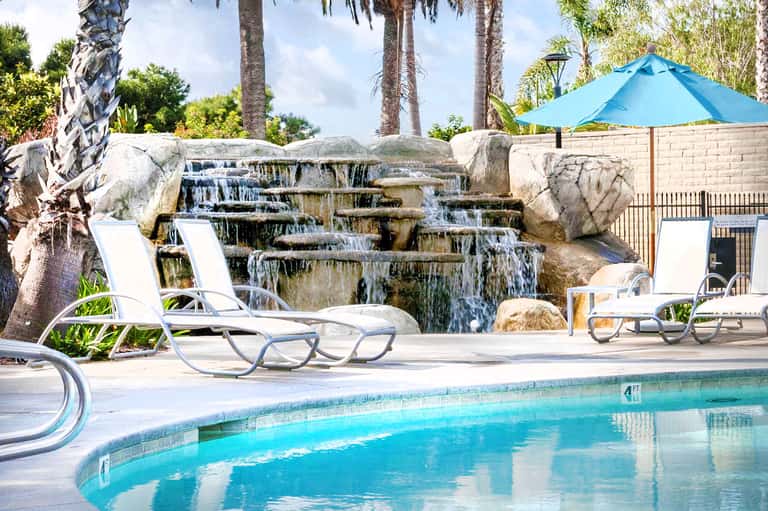 Resort pool with cascading waterfall, lounge chairs, palm trees, and blue umbrella on sunny day