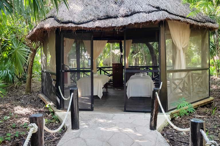 Thatched-roof spa pavilion with black frame, white curtains, massage tables, and tropical vegetation surroundings