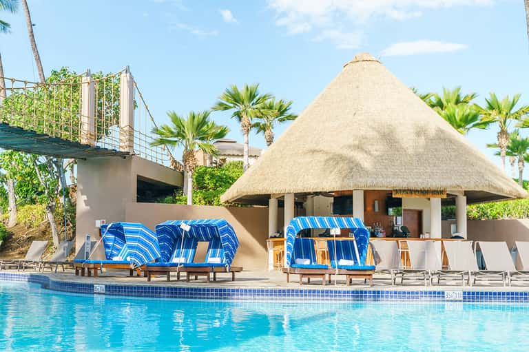 Resort pool area with thatched-roof cabana, blue loungers, and palm trees under clear sky
