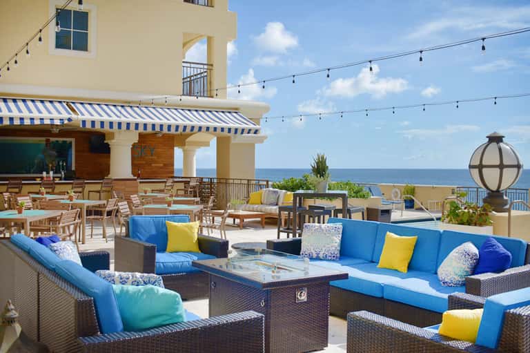 Oceanfront terrace with blue and yellow cushioned seating, striped awning, and string lights overlooking the sea