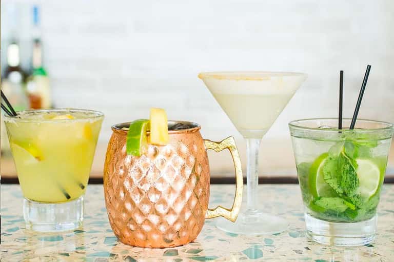 Four colorful cocktails on a table, including a Moscow Mule in a copper mug and a mojito with mint