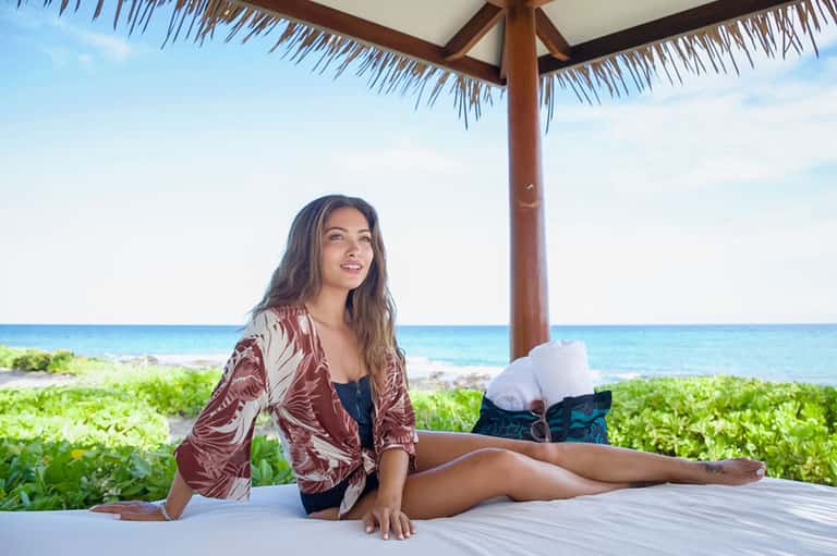 Woman in patterned jacket relaxing on beach platform under thatched umbrella with ocean view