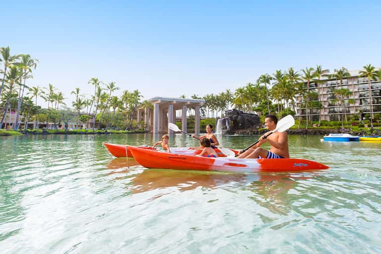 Family kayaking in shallow tropical waters near palm-lined resort with modern architecture