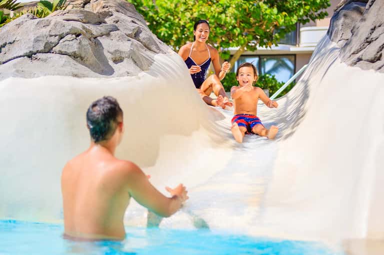 Family playing in shallow resort pool with large white boulder water slide feature