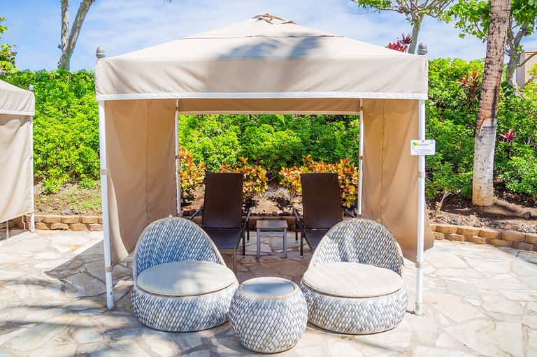 Two white woven lounge chairs under shaded cabana with flowering plants backdrop