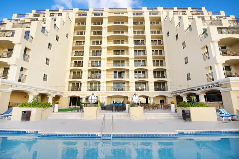 Beige multi-story resort building with curved balconies overlooking a rectangular swimming pool under blue sky