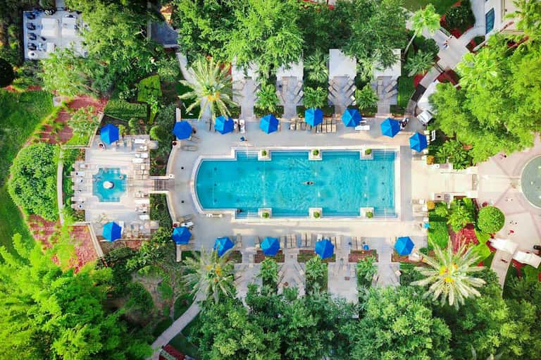Aerial view of resort pool surrounded by blue umbrellas, loungers, and lush green landscaping