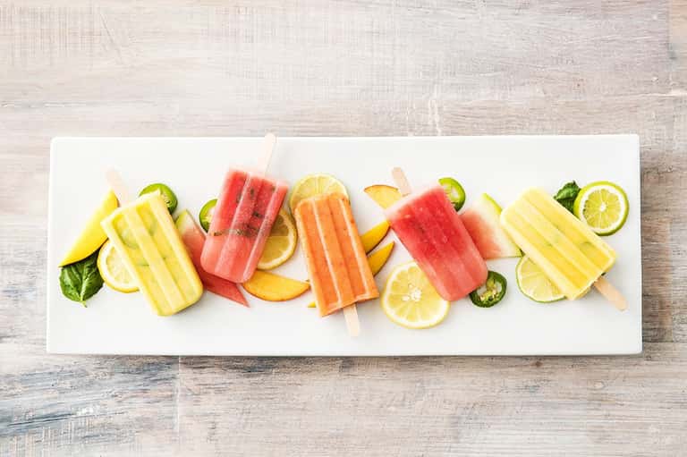 Colorful fruit popsicles and fresh fruit slices arranged on a white platter