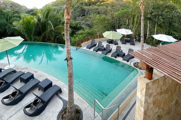 Turquoise infinity pool with black lounge chairs and wooden deck in lush jungle setting
