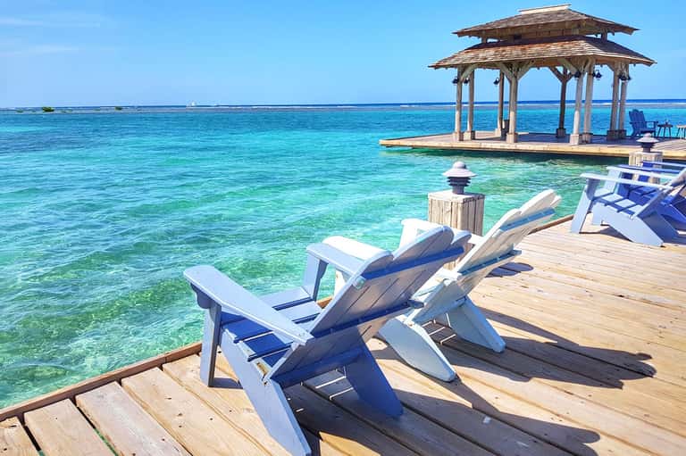 Wooden dock with white and blue Adirondack chairs facing crystal-clear turquoise water and gazebo