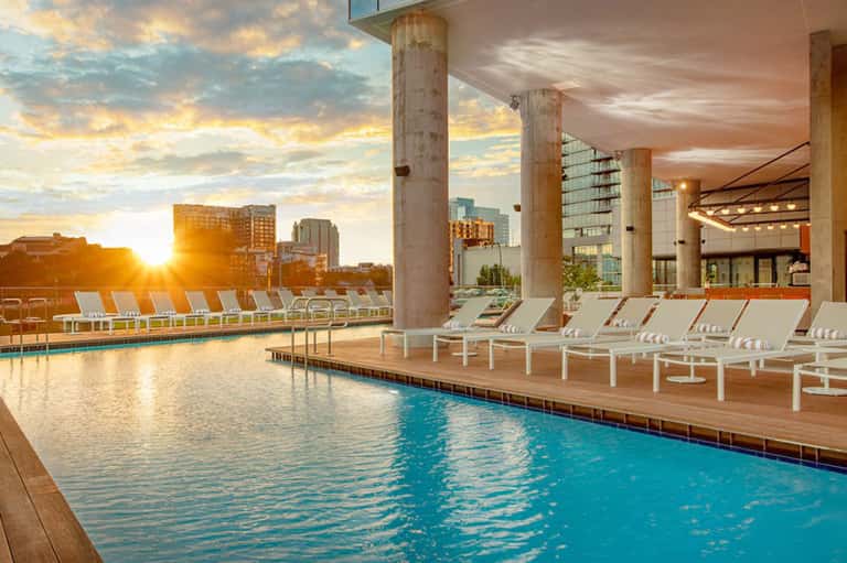 Indoor pool with natural light, white lounge chairs, and sunset city skyline views through large windows