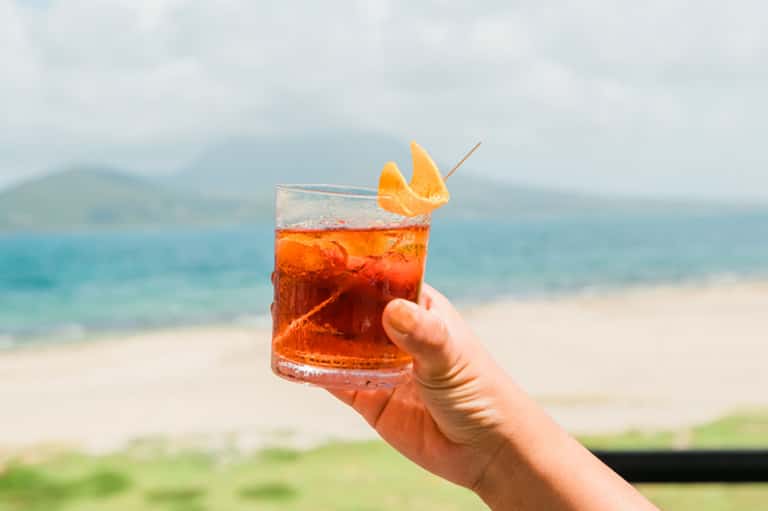 Hand holding a vibrant red cocktail with orange garnish against beach and ocean backdrop