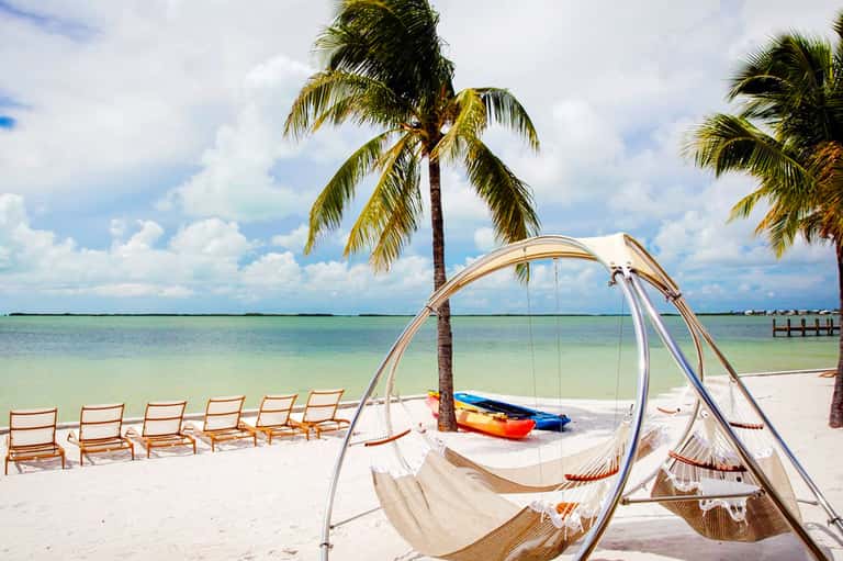 Tropical beach with hammocks, palm trees, kayaks, and turquoise water under blue sky