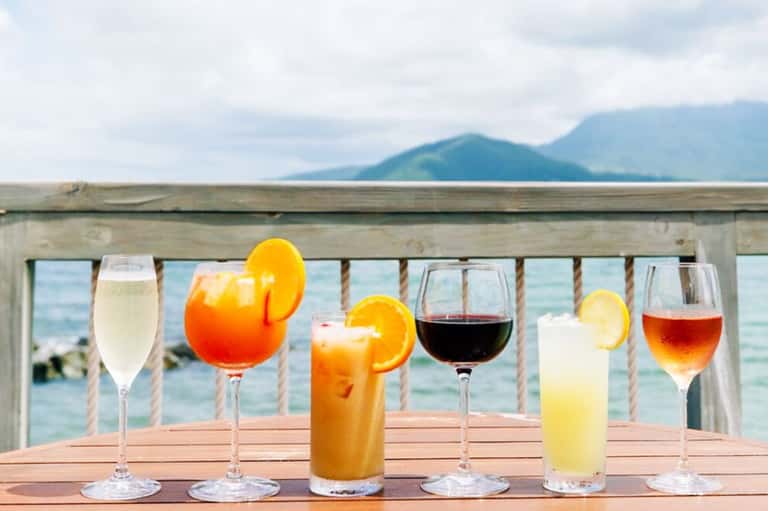 Six colorful tropical cocktails displayed on wooden waterfront railing with mountain views
