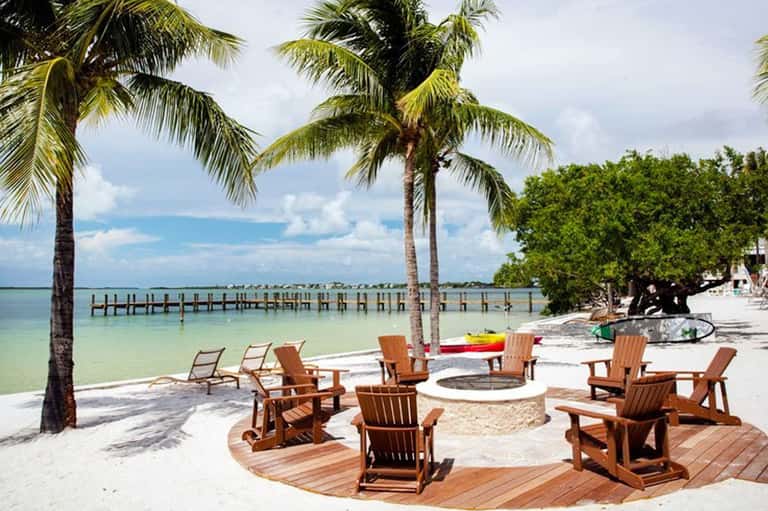 Beach resort with wooden Adirondack chairs around fire pit, palm trees, and wooden pier extending into turquoise water
