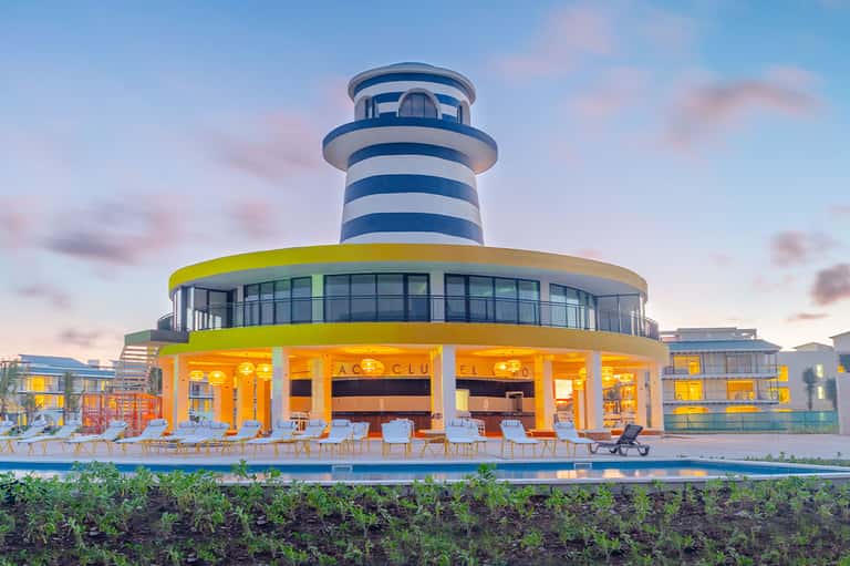Modern beachfront building with blue and white striped lighthouse tower and yellow-lit ground floor at dusk