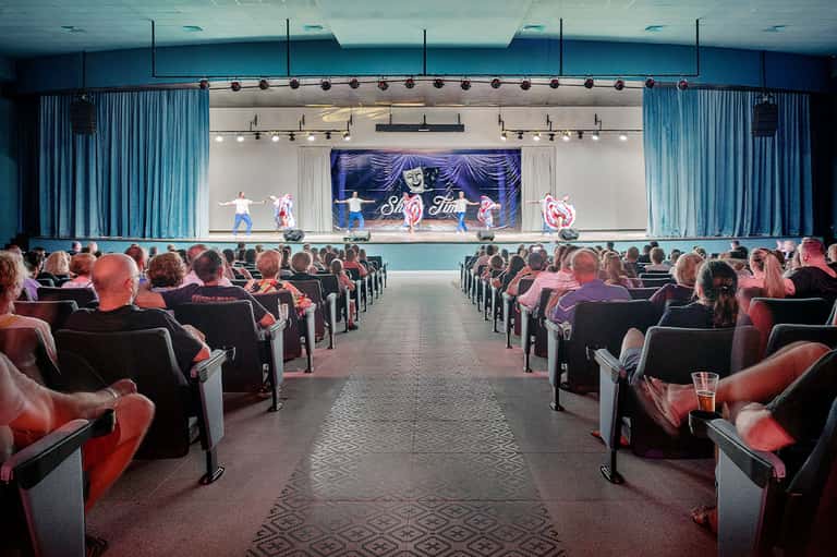 Theater auditorium with red seats, blue curtains, and stage performance with dancers and neon signage