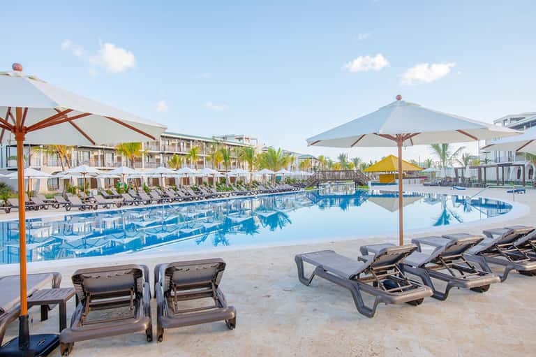 Resort pool with lounge chairs and white umbrellas surrounded by palm trees and modern architecture