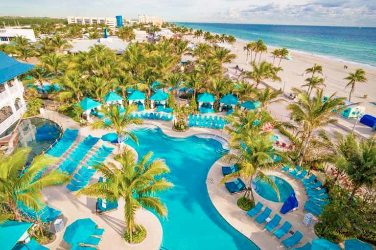Aerial view of beachfront resort with multiple turquoise pools, palm trees, and pristine sandy beach with ocean beyond