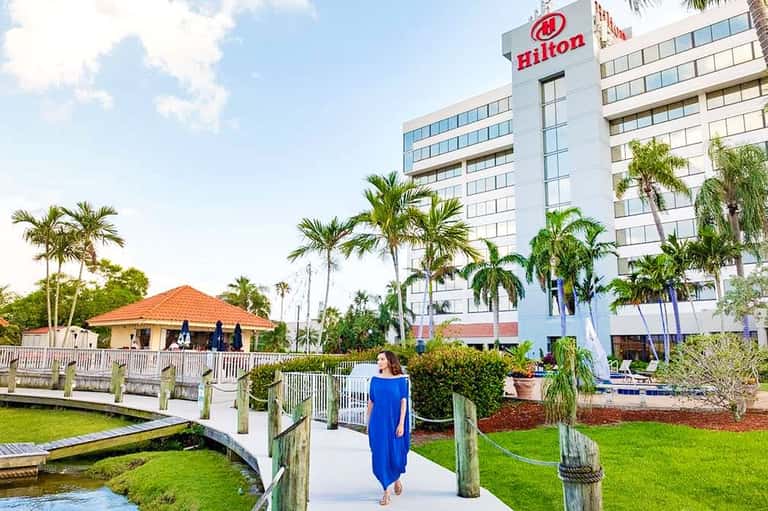 Hilton hotel exterior with grounds, wooden bridge, and woman in blue dress walking