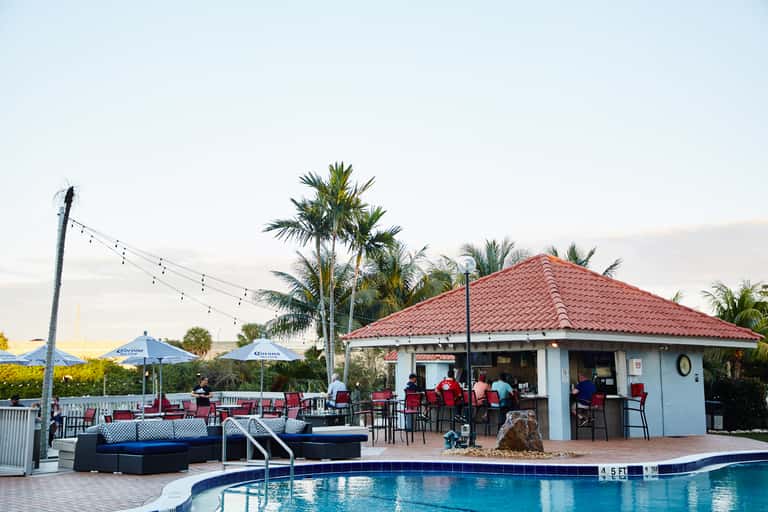 Poolside bar and lounge area with palm trees, red tile roof, and blue umbrellas