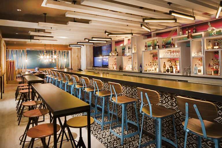 Modern bar interior with high seating, geometric patterns, pendant lighting, and shelved beverages
