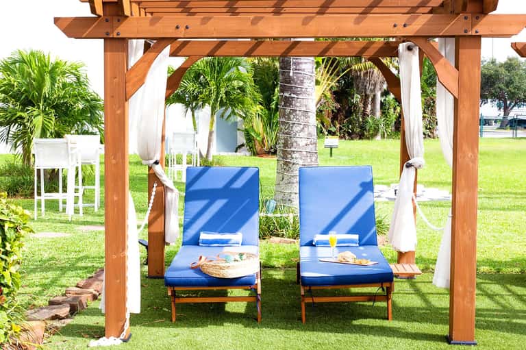 Wooden pergola over green lawn with two blue lounge chairs and tropical plants.