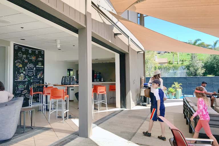 Modern outdoor patio with open kitchen, orange bar stools, shade sail, and family playing cornhole