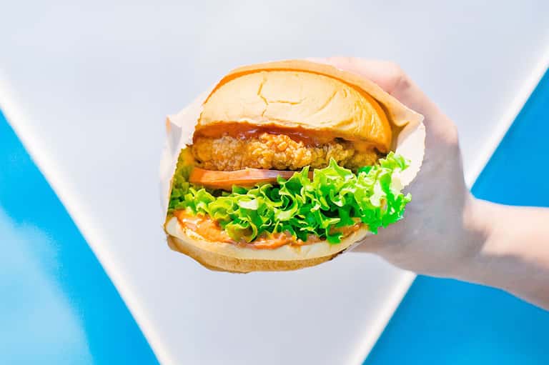 Gourmet burger with crispy patty, fresh lettuce, tomato, and golden bun held in hands against blue background