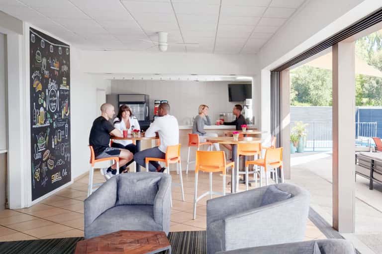 Modern hotel lounge with orange chairs, communal dining area, and outdoor terrace access