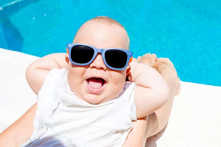 Happy baby wearing sunglasses by a bright blue swimming pool
