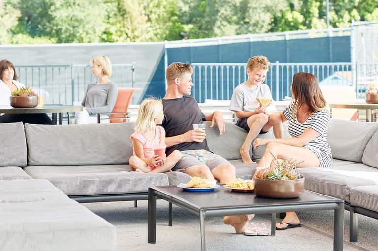 Family relaxing on modern outdoor sectional sofa by swimming pool with snacks and greenery