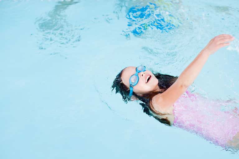Child swimming backstroke in bright blue pool water wearing pink swimsuit and goggles
