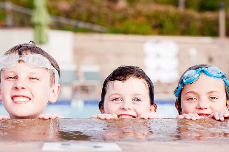 Three children in swimming goggles smiling in a resort pool
