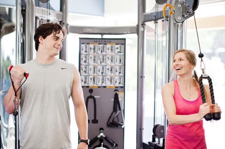 Young man and woman using cable exercise equipment in a modern gym with floor-to-ceiling windows